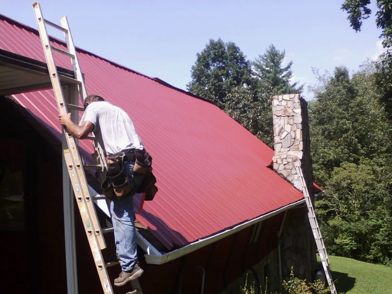 Roofer on a ladder beside a red metal roof and stone chimney — Soffit & Fascia Repair by Roofing Brothers in Green Mountain